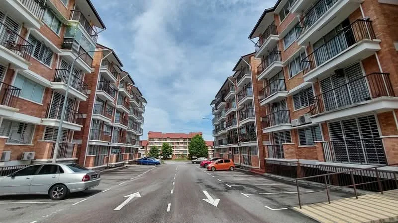 Exterior type: low-rise apartment block; features: red brick facade, balconies with black railings, multiple storeys, asphalt parking lot, marked parking spaces, surrounding greenery, clear blue sky, two-way street layout; style: modern.