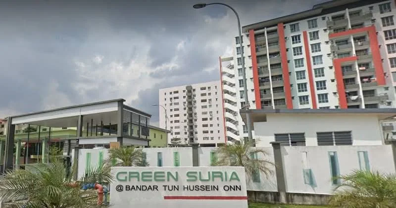 Exterior type: low-rise apartment block; features: white painted facade, red accent panels, glass curtain wall, manicured landscaping, palm trees, signage with 'GREEN SURIA', multiple storeys, balconies, parking area visible, surrounded by greenery; style: modern.