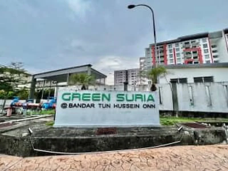 Exterior features: signage with 'GREEN SURIA', landscaped area, paved walkway, surrounding greenery, multi-storey building in background, parking area visible, street lamp, cloudy sky; style: modern.