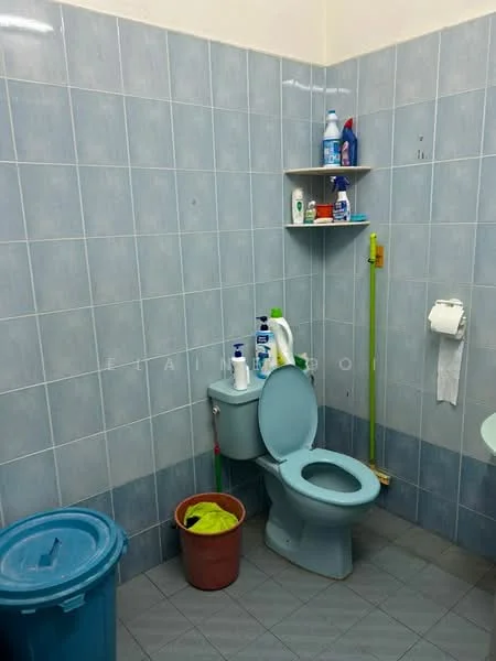 Bathroom features: blue tiled walls, grey tile flooring, one-piece toilet, toilet paper holder, shelf with cleaning supplies, green trash bin, red bucket, sink with faucet.