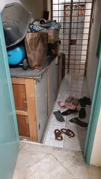 Room features: grey tiled flooring, wooden cabinetry, countertop with clutter, blue storage container, cardboard boxes, slippers, sandals, metal security grille door, white walls, light source from adjacent room.