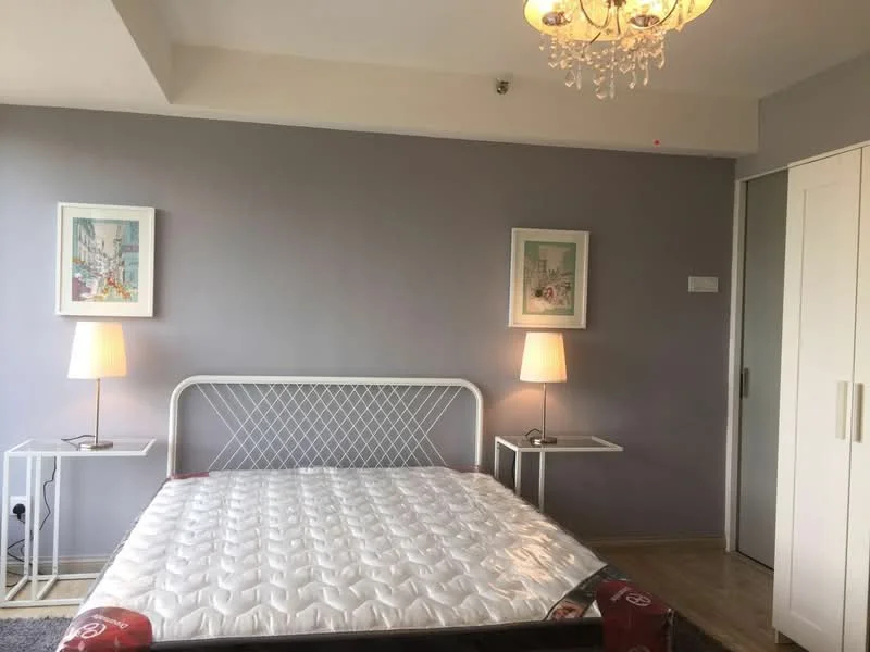 Bedroom features: queen-size bed, white metal bed frame, mattress with protective cover, two bedside tables, table lamps with white shades, light purple painted walls, wooden flooring, framed artwork on walls, built-in wardrobe with sliding doors, chandelier light fixture; style: modern.