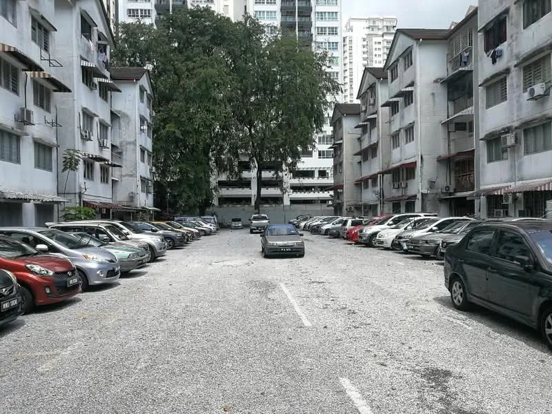 Exterior type: low-rise apartment block; features: gravel parking lot, multiple parked cars, surrounding low-rise buildings, mature trees, visible air conditioning units on buildings, open-air parking, two-storey buildings, balconies on upper floors; style: traditional.