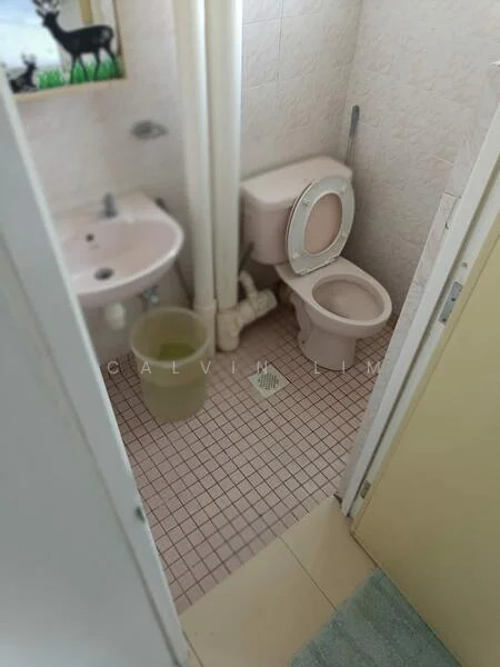 Bathroom features: pink ceramic sink, toilet, pink tiled walls, pink tiled flooring, shower drain, plumbing pipes visible, small waste bin, bath mat, mirror above sink.