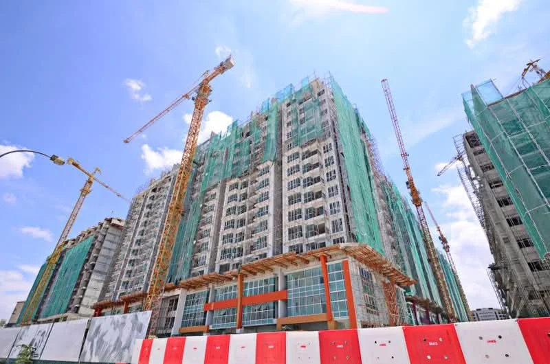 Exterior type: high-rise condominium; features: glass curtain wall, construction scaffolding, green construction netting, cranes on site, multiple storeys, exposed concrete structure, red accent panels, surrounding construction barriers; style: modern.