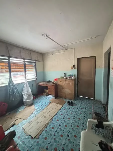 Room features: blue and white painted walls, concrete flooring with patterned design, wooden desk, wooden cabinet, metal door, window with wooden slats, plastic chair, cardboard on the floor, plastic bags, ceiling-mounted light fixture.