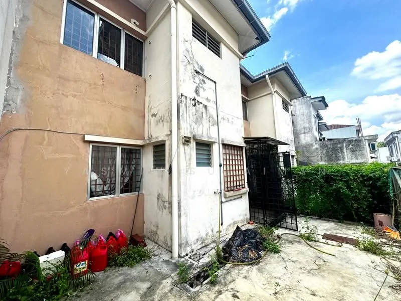 Exterior type: semi-detached house; features: two-storey structure, cream painted facade, exposed brick wall, metal security grille, concrete flooring, overgrown landscaping, visible plumbing pipes, backyard area, storage area with red containers, partially cloudy sky; style: modern.