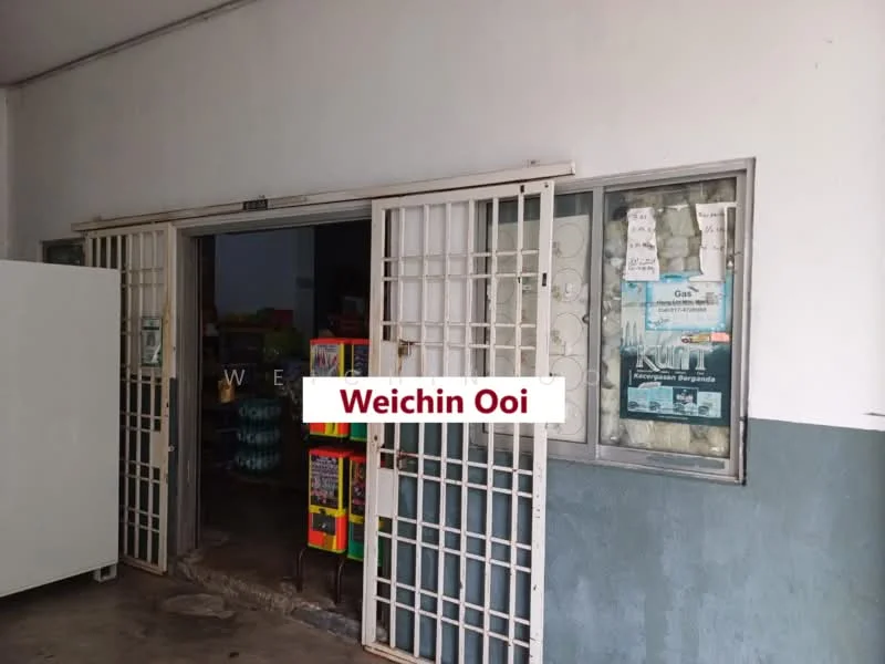 Exterior features: double-door entrance with security grills, glass window with signage, concrete walls with grey paint, flooring visible inside, shelves visible through the window, various colorful items displayed inside.