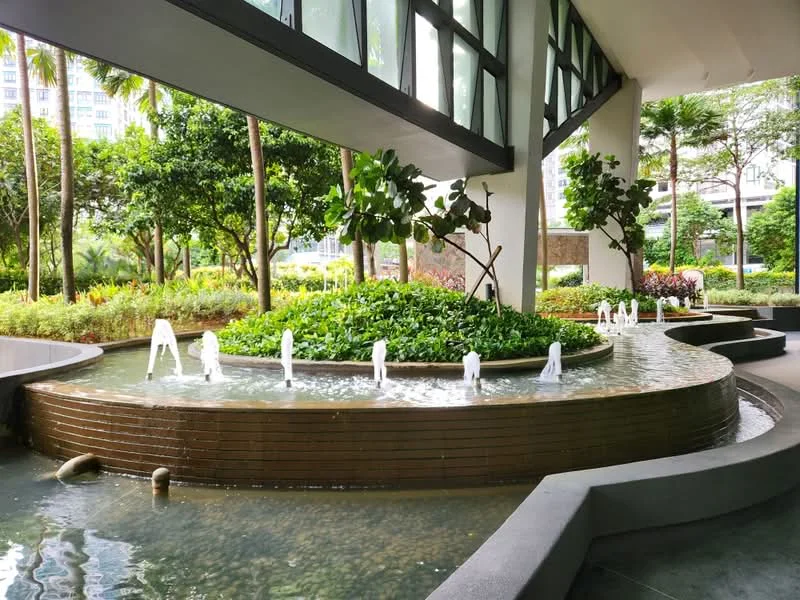 Exterior type: landscape garden; features: water feature with fountain jets, curved stone seating area, manicured garden beds, tropical landscaping with palm trees, surrounding greenery, pebble stone pathway, modern architectural elements, large glass windows; style: modern.