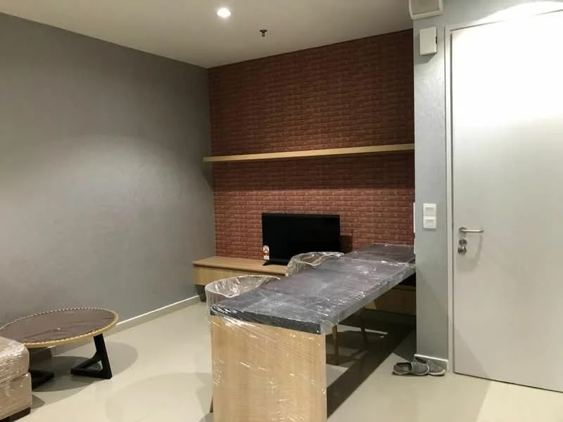 Living features: grey painted walls, exposed brick accent wall, light-colored flooring, wooden TV console, black television, glass-top coffee table, plastic-wrapped dining chairs, suspended wooden shelf, white door, recessed lighting; style: modern.