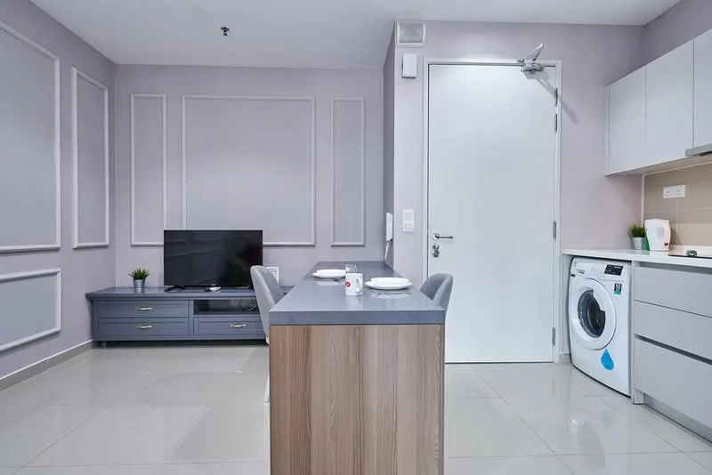 Kitchen features: grey cabinetry, white countertop, built-in washing machine, black flat-screen TV, dining table with two chairs, light grey walls with paneling, recessed lighting, ceramic tile flooring; style: modern.