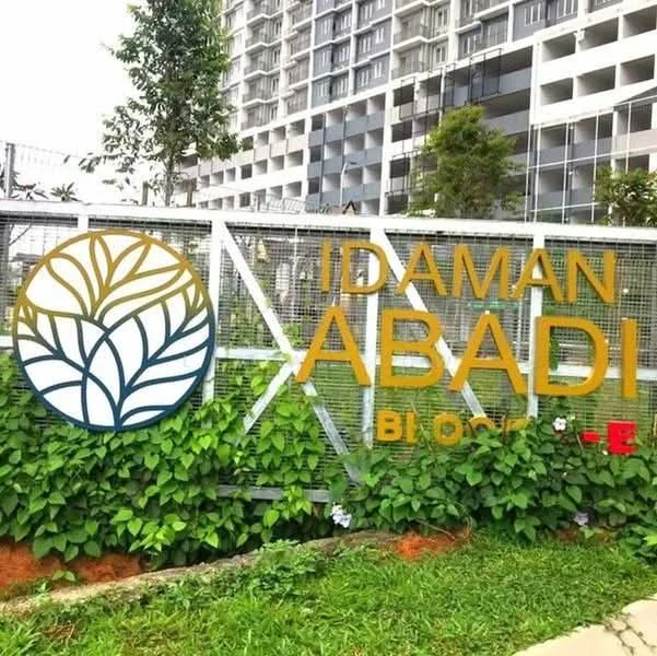 Exterior features: signage with 'IDAMAN ABADI' text, decorative leaf design on signage, greenery and plants surrounding the sign, modern building facade in background, multi-storey residential building, fencing around the property; style: modern.