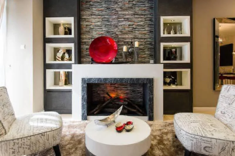 Living features: stone feature wall, white painted walls, fireplace with black surround, decorative red bowl on mantel, two accent chairs with printed upholstery, round coffee table, decorative silver bowl on table, candles on mantel, built-in shelves with decorative items, area rug, recessed lighting; style: modern.
