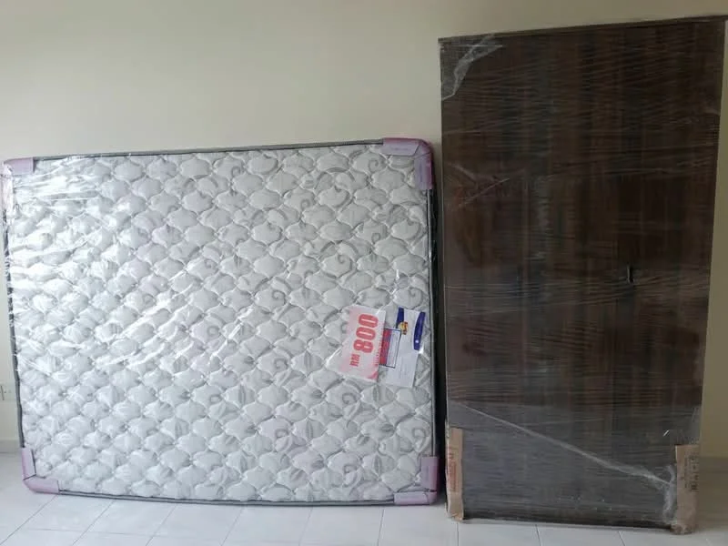 Room features: mattress with spring coils, wooden wardrobe, white tiled flooring, neutral wall color, plastic wrap on wardrobe.