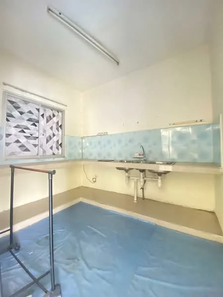 Kitchen features: white painted walls, blue tiled backsplash, single-bowl stainless-steel sink, chrome faucet, countertop with storage underneath, window with patterned treatment, flooring covered with blue tarp, overhead fluorescent lighting.