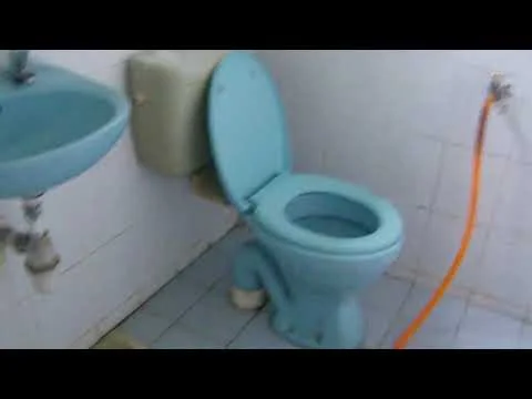 Bathroom features: blue toilet, blue sink, white tiled walls, blue tiled flooring, exposed plumbing, orange water hose, wall-mounted faucet, mirror above sink.