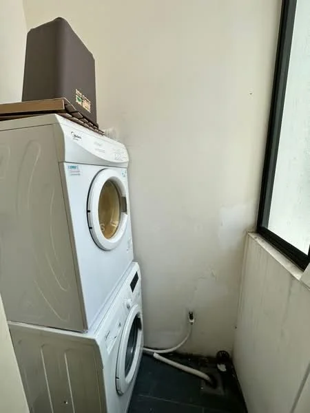 Laundry features: front-load washing machine, front-load dryer, white appliance color, tile flooring, window with natural light, wall with peeling paint, plumbing connections visible, stacked washer and dryer.