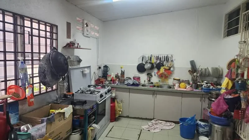 Kitchen features: white painted walls, tile flooring, stainless-steel gas stove, stainless-steel sink, grey laminate countertop, wall-mounted shelves, various cooking utensils, multiple pots and pans, plastic storage containers, kitchen towels, open shelving with dishes, window with metal grille, red fire extinguisher, blue plastic bucket.
