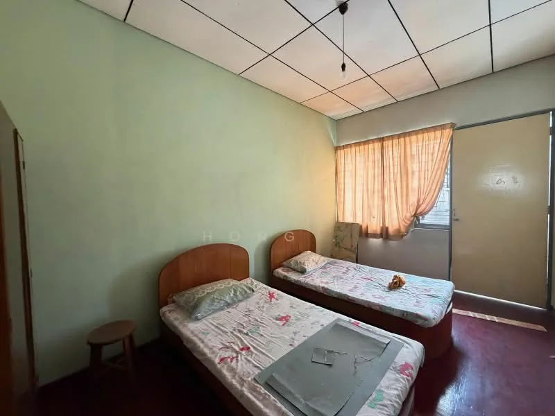 Bedroom features: two single beds with wooden headboards, floral-patterned bed linens, wooden stool, green painted walls, brown wooden flooring, curtains in light orange, window with natural light, ceiling light fixture.