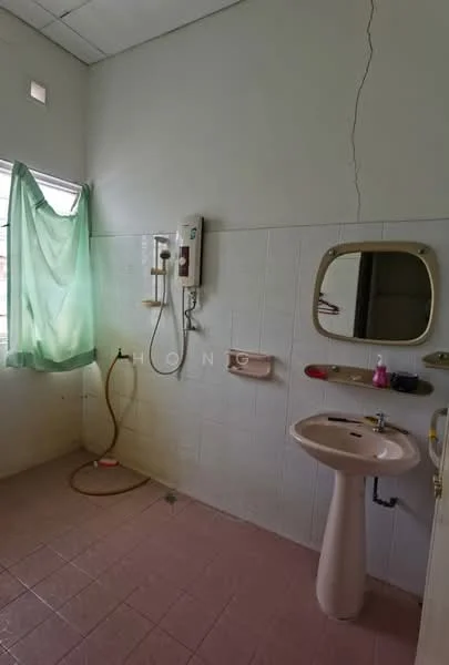 Bathroom features: white tiled walls, pink tiled flooring, single-sink pedestal vanity, chrome basin mixer faucet, framed mirror, shower area with wall-mounted shower head, shower hose, toilet paper holder, window with sheer green curtain, exposed plumbing, light fixture, wall-mounted soap dish.