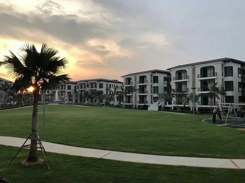Exterior type: low-rise apartment block; features: green lawn, palm trees, playground equipment, walking path, balconies on buildings, landscaped garden, sunset view, manicured landscaping; style: modern.