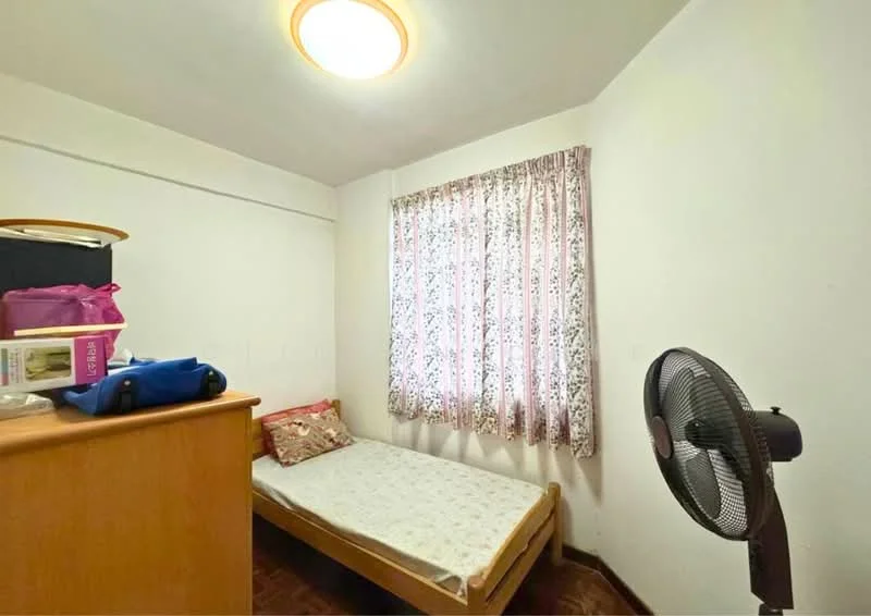 Bedroom features: single bed, wooden bed frame, floral-patterned bedspread, wooden dresser, table fan, curtains with floral pattern, white painted walls, laminate flooring, ceiling light fixture.