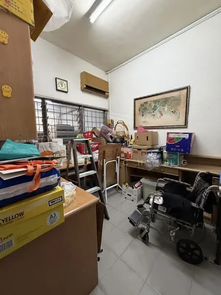 Room features: white painted walls, grey tiled flooring, window with metal grilles, wall-mounted air conditioning unit, ceiling lights, wooden shelving unit, cardboard boxes, folding chairs, wheelchair, framed artwork, clock on the wall.