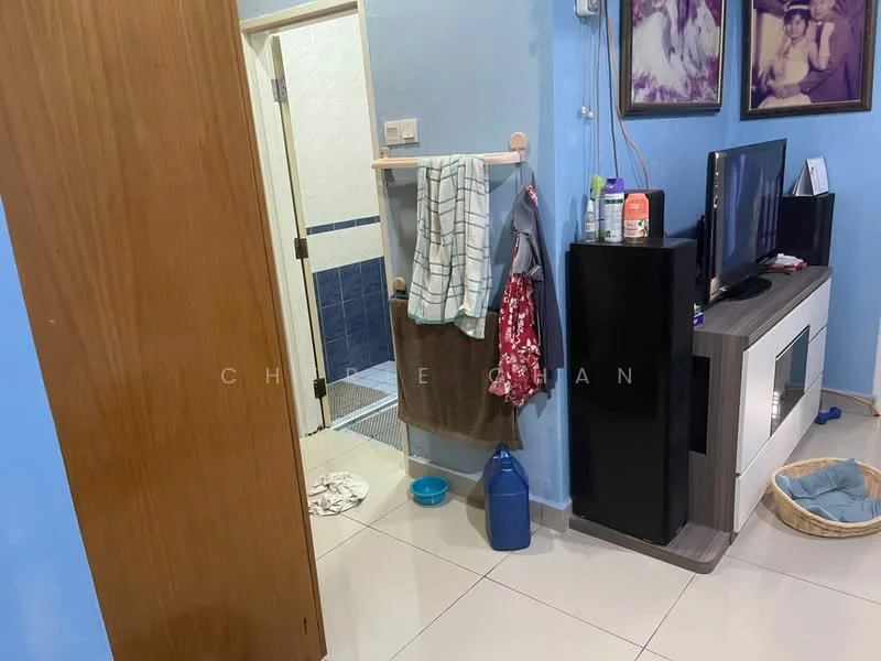 Room features: blue painted walls, white tiled flooring, wooden door, wall-mounted TV, black TV console, towel rack, hanging towels, cleaning supplies on console, laundry basket, bathroom door, shower area visible, white bath mat.