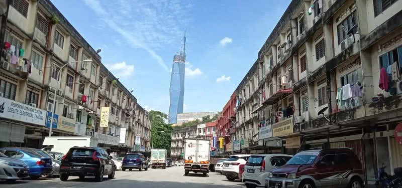 Exterior type: shophouse; features: multi-storey shophouse, ground floor retail spaces, residential units above, balconies with railings, visible laundry hanging, street view with parked cars, urban context with nearby buildings, KL skyline in background, Petronas Twin Towers visible, paved street, clear blue sky; style: traditional.