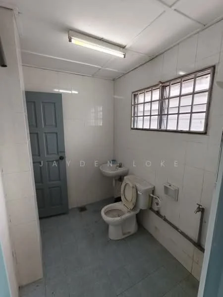 Bathroom features: white tiled walls, light blue tiled flooring, single-sink vanity with wall-mounted faucet, one-piece toilet, window with metal grille, overhead fluorescent lighting, grey door, exposed plumbing, mirror above sink.