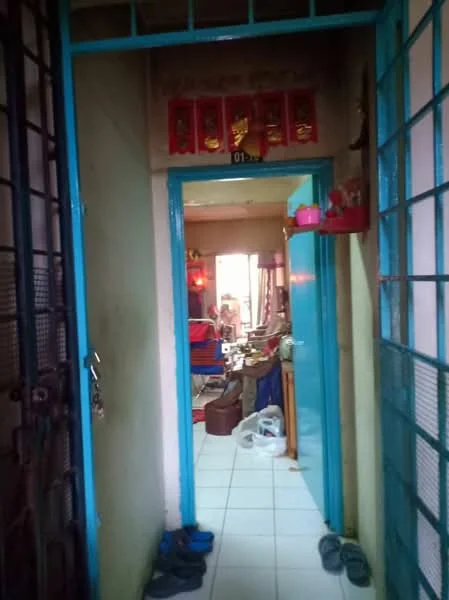 Room features: blue painted walls, white tiled flooring, wooden door, metal security gate, decorative wall hangings, visible furniture in background, plastic bags on floor, slippers on floor.