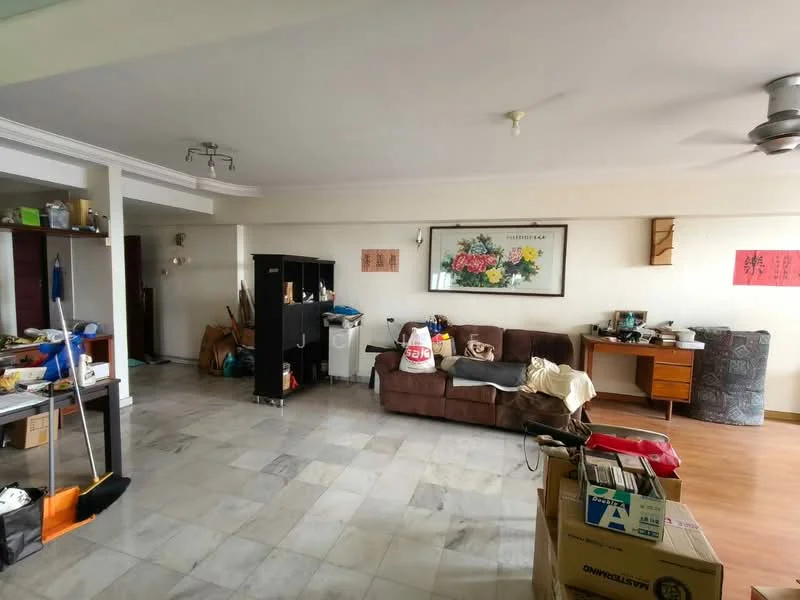 Living features: grey marble flooring, white painted walls, 5-blade ceiling fan, sofa in brown fabric, wooden side table, framed artwork on wall, open shelving unit, cardboard boxes, cleaning supplies, desk with chair, decorative items on shelves.
