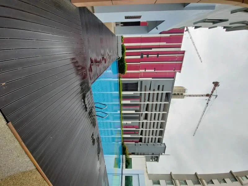 Exterior type: high-rise condominium; features: infinity pool, pool deck with wooden flooring, surrounding landscaping with greenery, glass railing, modern architectural design, construction cranes in background, multi-storey building facade with pink and grey elements; style: modern.