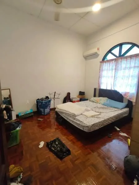 Bedroom features: wooden flooring, white painted walls, ceiling fan, window with sheer curtains, double bed with mattress, mirror, laundry basket, clothes rack, scattered items on the floor, air conditioning unit.