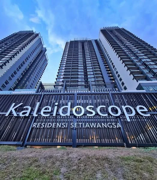 Exterior type: high-rise condominium; features: glass curtain wall, metal cladding, recessed balconies, feature lighting at night, landscaped entrance, signage with name 'Kaleidoscope', surrounded by greenery; style: modern.