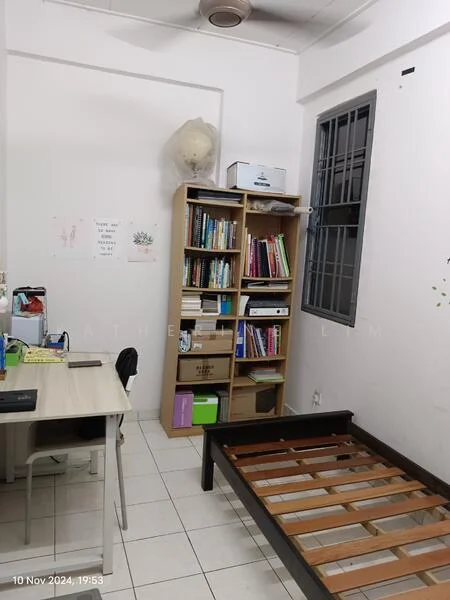 Room features: white painted walls, tile flooring, single bed frame without mattress, wooden bookshelf with books, desk with chair, window with metal grille, ceiling fan, decorative wall art, storage boxes, greenery in pots; style: minimalist.