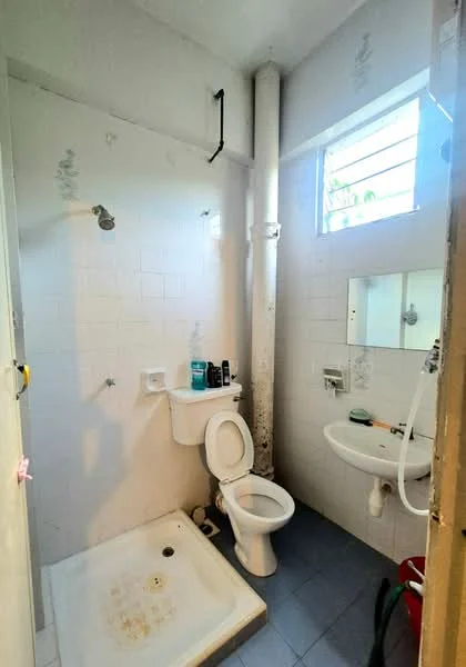 Bathroom features: white tiled walls, blue ceramic tile flooring, shower area with shower head, toilet, sink with mirror, wall-mounted faucet, window for natural light, plumbing pipe visible.