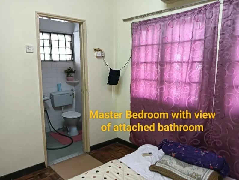 Bedroom features: cream painted walls, wooden flooring, purple sheer curtains, double bed with bedding, decorative pillows, window with grid pattern, wall-mounted light switch, door leading to bathroom. Bathroom features: white tiled walls, toilet, sink, shower hose, small potted plant, window for ventilation.