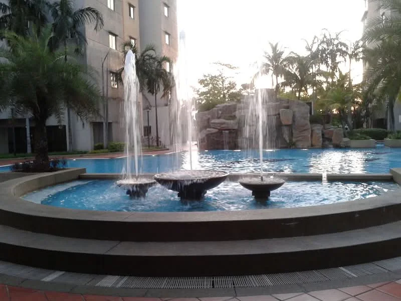 Exterior type: swimming pool deck; features: swimming pool with fountain, rock feature in pool area, palm trees surrounding pool, manicured landscaping, concrete pool deck, water feature with multiple jets, seating area around pool, adjacent building facade, evening lighting, tropical landscaping; style: modern.