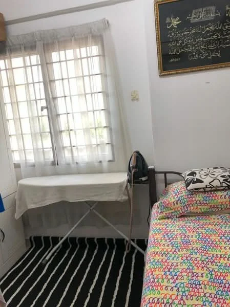 Bedroom features: single bed with colorful bedding, ironing board, window with white curtains, striped area rug, wall-mounted artwork, wooden bed frame, painted walls, light-colored ceiling.