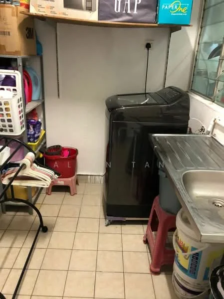 Laundry features: front-load washing machine in black, stainless-steel sink, grey countertop, tile flooring, white painted walls, plastic laundry basket, storage boxes, cleaning supplies, plastic stool, shelving unit.