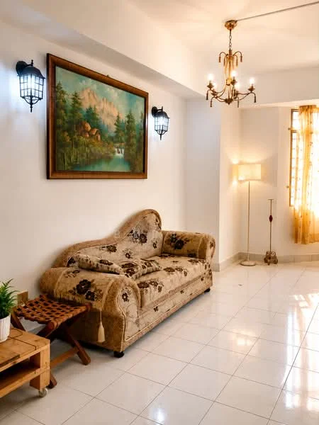 Living features: 3-seater fabric sofa in beige with floral pattern, framed landscape painting on wall, 2 wall-mounted lantern-style sconces, chandelier with multiple bulbs, floor lamp with fabric shade, white painted walls, glossy white tile flooring, wooden side table, small wooden stool, curtains in light yellow, natural light from window; style: traditional.