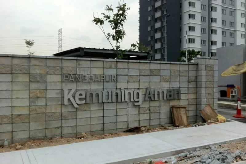 Exterior features: stone-clad wall, signage with 'Kemuning Aman', surrounding landscaping with small trees, paved walkway, construction materials visible, residential building in background; style: modern.