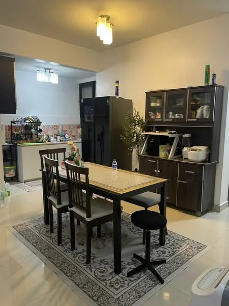 Dining features: dining table with beige tabletop, 4 dining chairs with black upholstery, area rug with decorative pattern, black wooden cabinet, refrigerator in black, kitchen counter visible in background, shelves with kitchenware, pendant lighting, white tiled flooring, green plant in pot; style: modern. Kitchen features: kitchen counter with tiled backsplash, various kitchen appliances visible, open shelving with kitchen items; style: modern.
