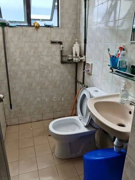 Bathroom features: white ceramic toilet, single-sink vanity with cream countertop, chrome basin mixer faucet, mirror above sink, tile flooring, grey tiled walls, shelf with toiletries, window for natural light, plumbing pipes visible, blue plastic container.