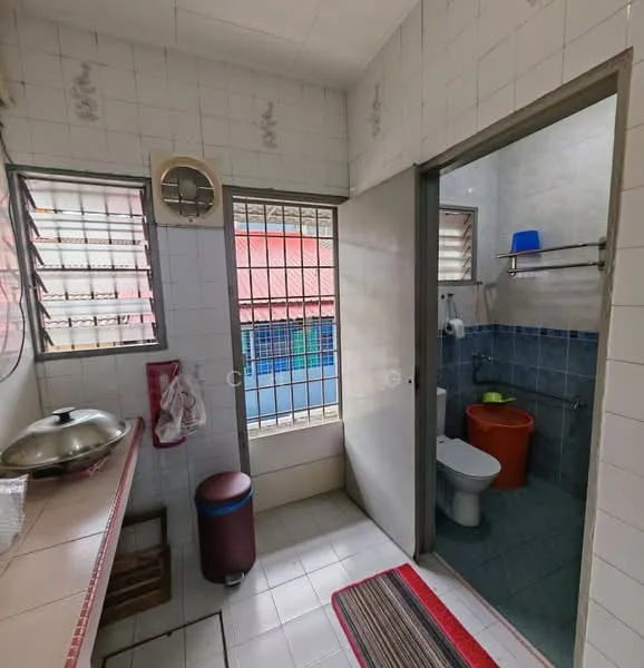 Room features: white tiled walls, light grey tiled flooring, single bowl stainless-steel sink, wooden countertop, window with metal bars, door leading to another room, toilet, orange bucket, shelf with metal rail, rug with stripes; style: functional.
