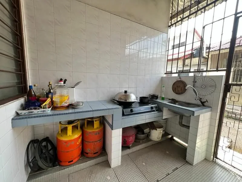 Kitchen features: white tiled walls, grey tiled countertop, single-bowl stainless-steel sink, chrome faucet, gas stove, frying pan, various cooking utensils, two gas cylinders, open shelving, floor tiles, window with metal grille.