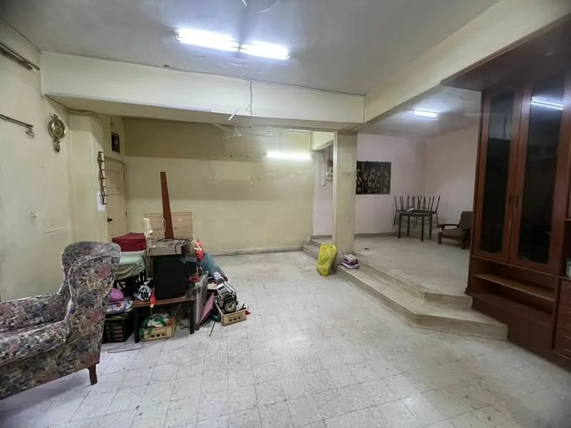 Room features: light-colored walls, tile flooring, fluorescent ceiling lights, steps leading to another area, furniture including a floral-patterned armchair, various boxes and items stored, empty wall space, open layout.