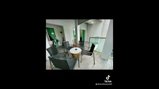 Room features: outdoor seating area with six black metal chairs, round white marble table, glass railing, grey tiled flooring, green curtains, exposed ceiling beams, natural light from large windows, decorative wall with textured finish; style: modern.