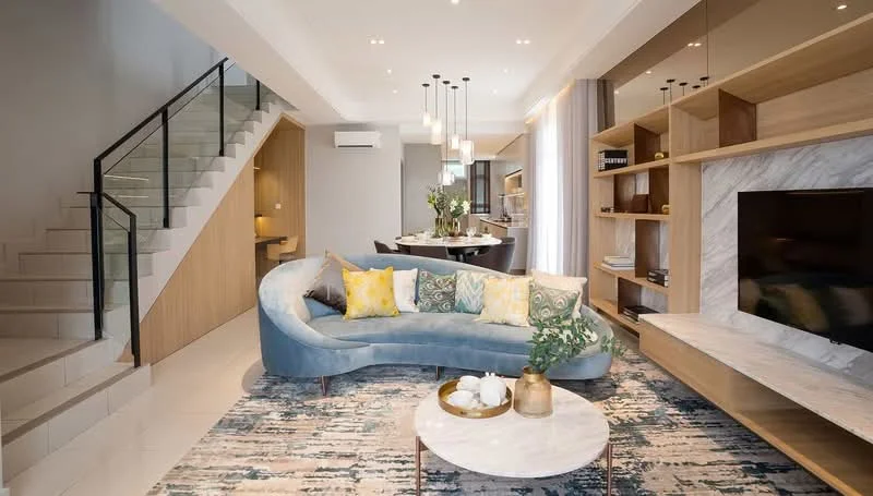 Living features: curved blue fabric sofa, yellow decorative cushions, marble coffee table, area rug with geometric pattern, wall-mounted TV, wooden TV console, built-in shelving unit, recessed ceiling lights, pendant lighting, floor-to-ceiling windows with sheer curtains, open-plan layout to dining area, staircase with wooden railing; style: modern.